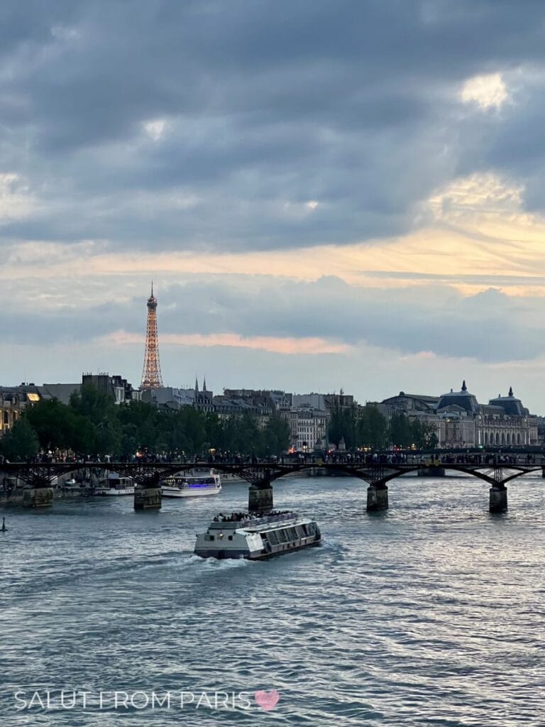 A sightseeing boat moves along the Seine at sunset with warm light reflecting on the water, while the Eiffel Tower glows in the distance behind a busy pedestrian bridge. The golden sky and iconic view highlight why this is a standout moment on the best Seine cruise in Paris. If you are looking for a seine dinner cruise paris, this will be your view.