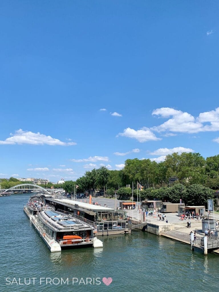A large river cruise boat is docked beside a lively quay where people gather near the water, with a modern bridge and leafy trees under a bright blue sky. This bustling boarding area shows the starting point for many of the best Seine cruise in Paris experiences.