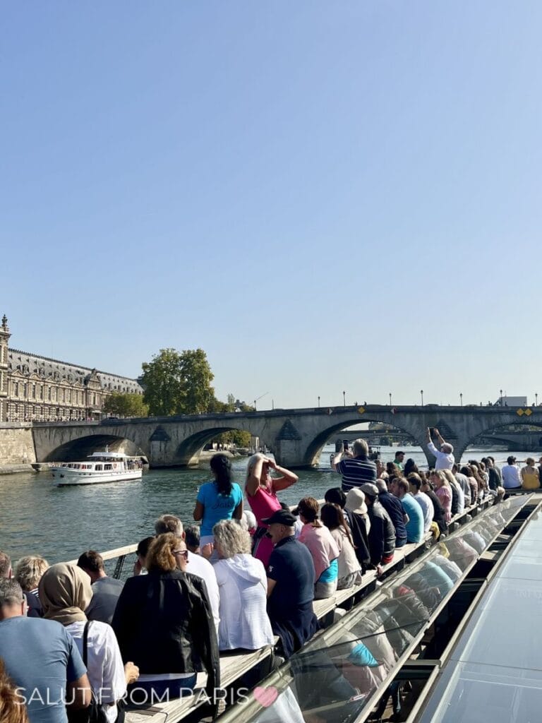 Tourists sit closely together on the open-air deck of a river cruise boat, many raising phones and cameras as they pass beneath a stone bridge along the Seine. The immersive viewpoint shows what it feels like to experience the best Seine cruise in Paris.
