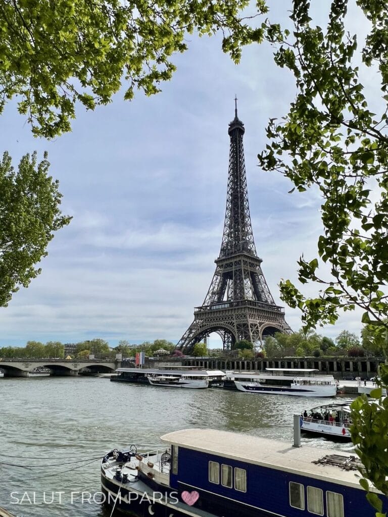 The Eiffel Tower stands tall beyond the river, framed by leafy green trees, while boats glide across the Seine in the foreground. This classic Paris view is one of the most memorable sights during the best Seine cruise in Paris.
