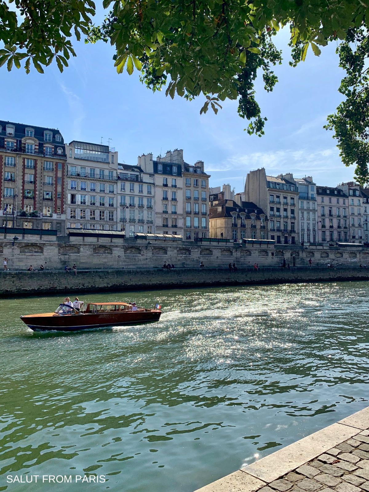 A small wooden boat glides along the Seine River in Paris in April as sunlight sparkles on the water. Classic Parisian buildings line the riverbank under a clear blue sky while leafy branches frame the scene above, with the text "SALUT FROM PARIS" visible in the corner.