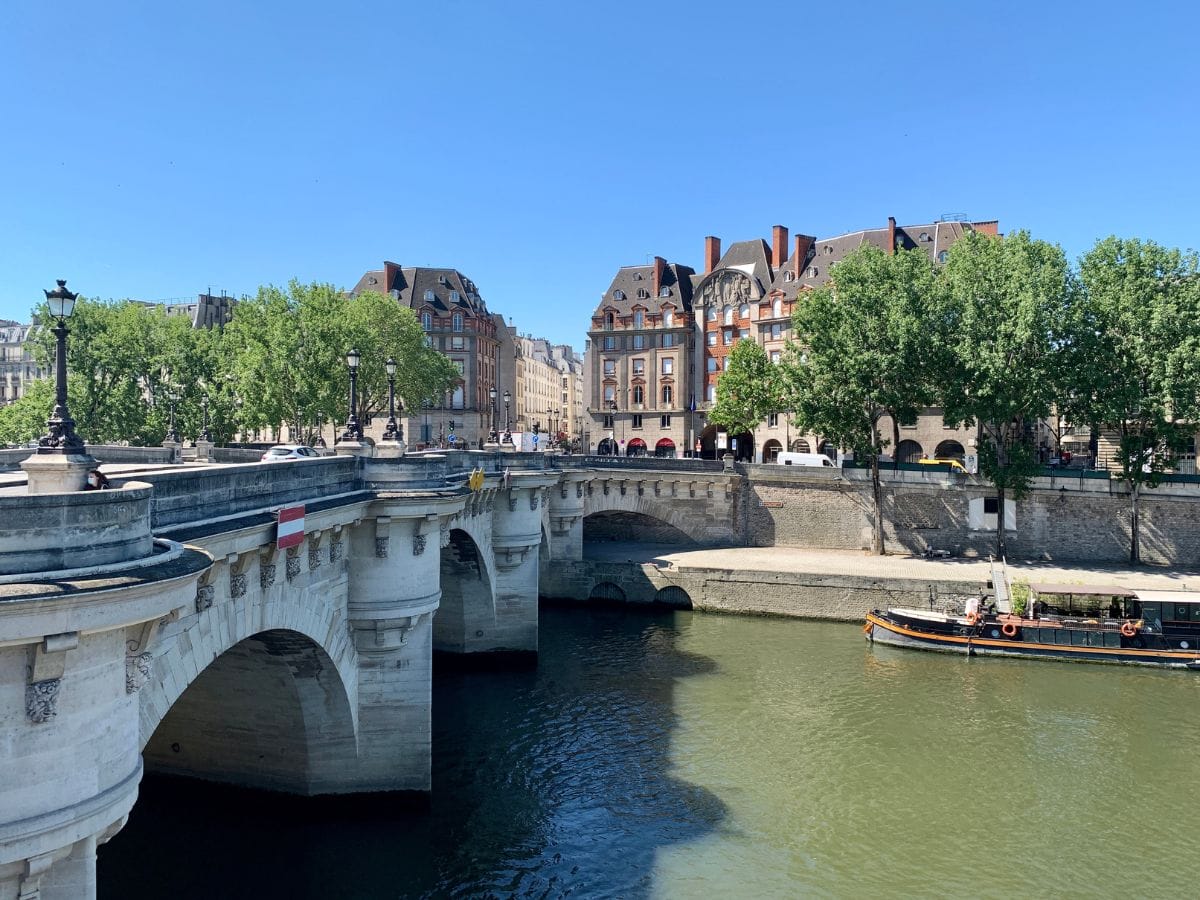 Stone bridge over the Seine with historic Paris buildings and trees lining the riverbank under clear blue skies showing a scenic riverside view in June in Paris.