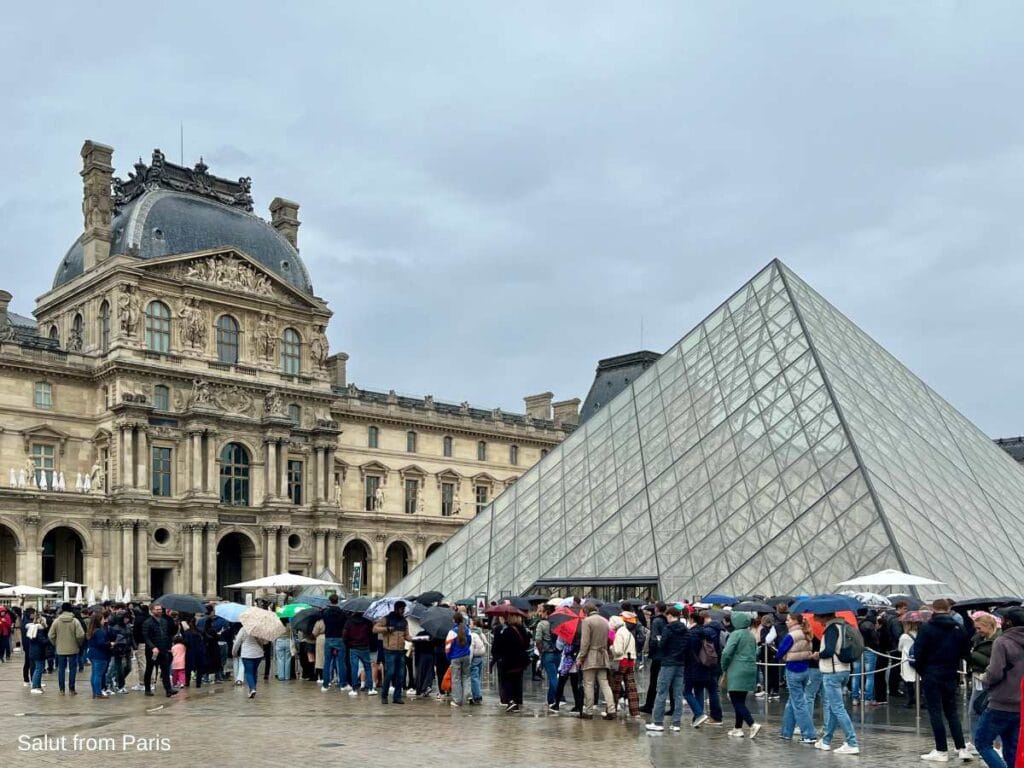 The image depicts a rainy day at the Louvre Museum in Paris, with a long line of people waiting under umbrellas in front of the iconic glass pyramid entrance. The historic Louvre building is visible in the background, showcasing its ornate architecture. The atmosphere is lively despite the overcast weather, capturing the bustling tourist activity around this famous landmark. In the lower left corner, the text "Salut from Paris" is visible, adding a postcard-like touch to the scene.
The image depicts a rainy day at the Louvre Museum in Paris, with a long line of people waiting under umbrellas in front of the iconic glass pyramid entrance. The historic Louvre building is visible in the background, showcasing its ornate architecture. The atmosphere is lively despite the overcast weather, capturing the bustling tourist activity around this famous landmark.