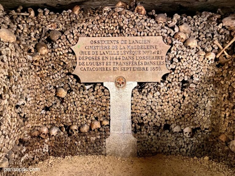 This image captures a haunting yet historically significant scene from the Catacombs of Paris, where human skulls and bones are meticulously stacked to form walls. A stone plaque in the center reads: "OSSEMENTS DE L'ANCIEN CIMETIรRE DE LA MAGDELEINE (RUE DE LA VILLE L'รVรQUE NOS 1 ET 2) DรPOSรS EN 1844 DANS L'OSSUAIRE DE L'OUEST ET TRANSFรRรS DANS LES CATACOMBES EN SEPTEMBRE 1859." This inscription indicates that these remains originated from the Magdeleine Cemetery and were moved to the ossuary in 1844, before being transferred to the Catacombs in 1859. The eerie ambiance, dim lighting, and tightly packed bones highlight the macabre beauty and historical depth of this underground burial site, which serves as a fascinating yet somber attraction in Paris.