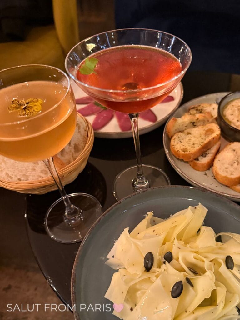 Two cocktails served alongside shared plates of thinly shaved cheese with seeds and toasted bread. The photo reads "SALUT FROM PARIS".