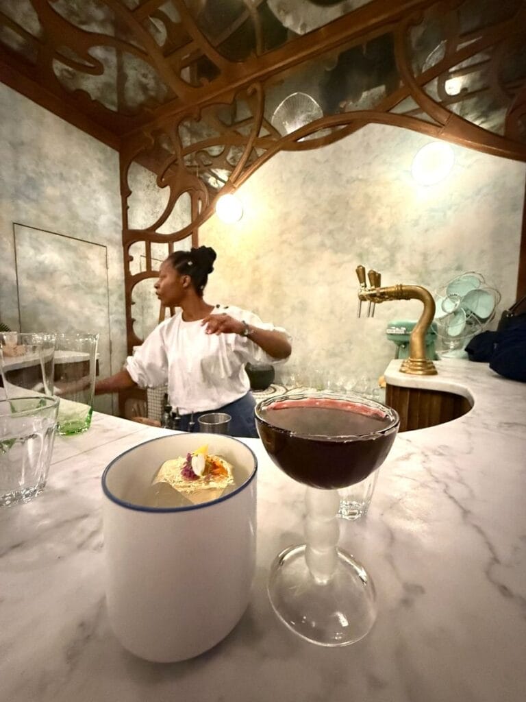 Dark red cocktail and a small white cup with a delicate edible garnish sit on a curved marble bar. A bartender works behind an ornate wood framed bar with brass taps.