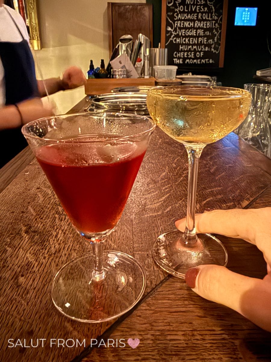 Two stemmed cocktails on a wooden bar Cambridge Public House including a deep red drink and a pale sparkling drink held by a hand. A handwritten menu is visible behind the bar and the photo reads "SALUT FROM PARIS".
