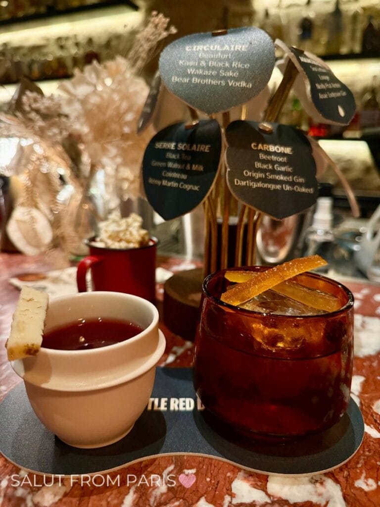 Two cocktails served on a decorative tray including a dark drink over a large ice cube with an orange peel and a warm drink in a white cup with a garnish on the rim. Cocktail ingredient cards behind the drinks read "CIRCULAIRE. Red Vermouth. Kushi and Black Rice Wakaze Sake. Bear Brothers Vodka." "SUCRE SOLAIRE. Black Tea. Green Walnut and Miso. Citron. Remy Martin Cognac." and "CARBONE. Beetroot. Black Garlic. Origin Smoked Gin. Dartigalongue Un Oaked." The photo reads "SALUT FROM PARIS".