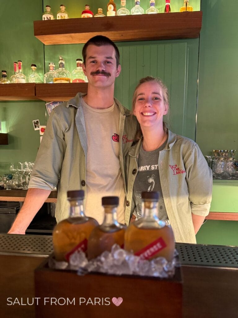 Two smiling bartenders stand together behind a green cocktail bar with liquor bottles displayed on wooden shelves. Bottles chill on ice in the foreground and the photo reads "SALUT FROM PARIS".