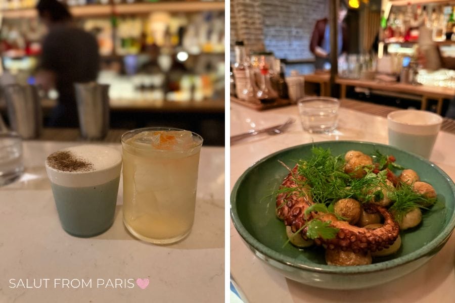 Two cocktail on a white table. The left one is in a turquoise ceramic cup with white foam and a darker powder, and the left is in a glass with icecubes and a orange powder on top. One the right is a plte with octpus and potatoes with green herbs as garnish.
