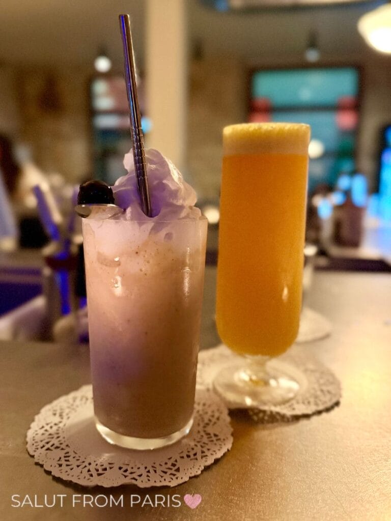 Brunch menue at Superfine: P&B Milkshake Two colorful cocktails at a Paris bar including a creamy purple drink topped with foam and a dark garnish and a tall frothy orange drink. The photo reads "SALUT FROM PARIS".