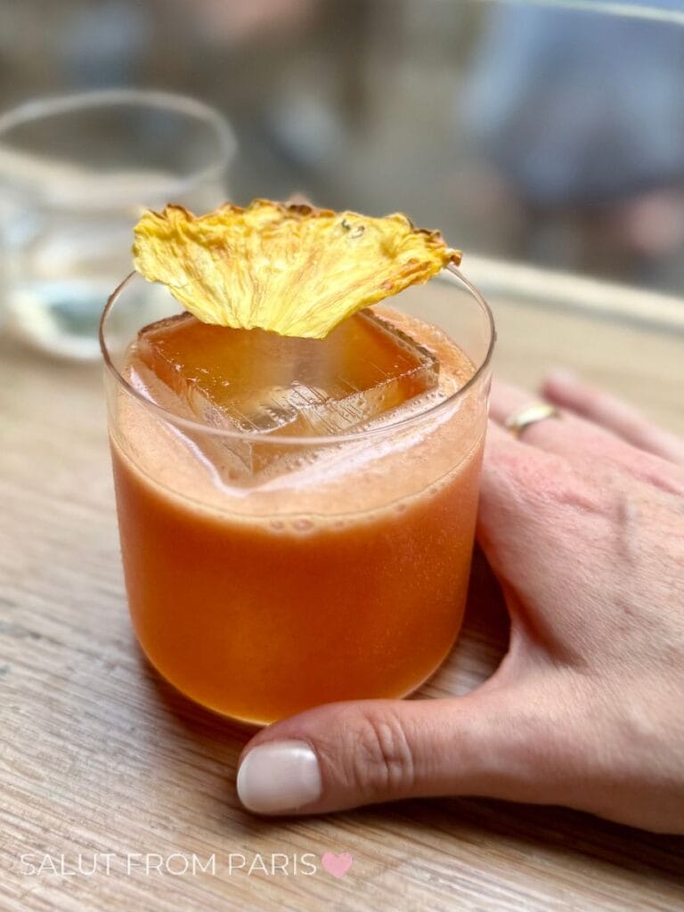 A hand holds an amber colored cocktail served over a large ice cube and topped with a dried pineapple garnish. The close up highlights the rich color and elegant presentation on a wooden table. Cozy cocktail bars are perfect for relaxing in Paris in Autumn.