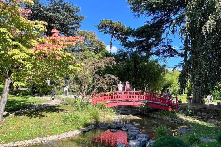 A serene scene from the Albert Kahn Museum and Gardens in Paris, featuring a vibrant red wooden bridge arching over a gently flowing stream. Lush greenery, including trees with hints of autumn colors, frames the tranquil Japanese-inspired landscape. A few visitors in light clothing stroll across the bridge, enjoying the peaceful surroundings under a bright blue sky.