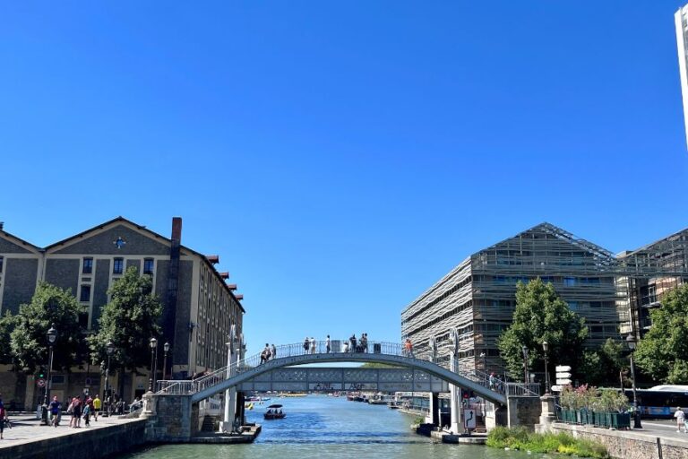This image captures a scenic view of a pedestrian bridge arching over the Canal Saint-Martin in Paris on a bright, sunny day. People are seen walking across the bridge, while boats glide through the calm waters below. The surrounding architecture blends modern glass buildings with traditional stone facades, framed by lush green trees against a clear blue sky.