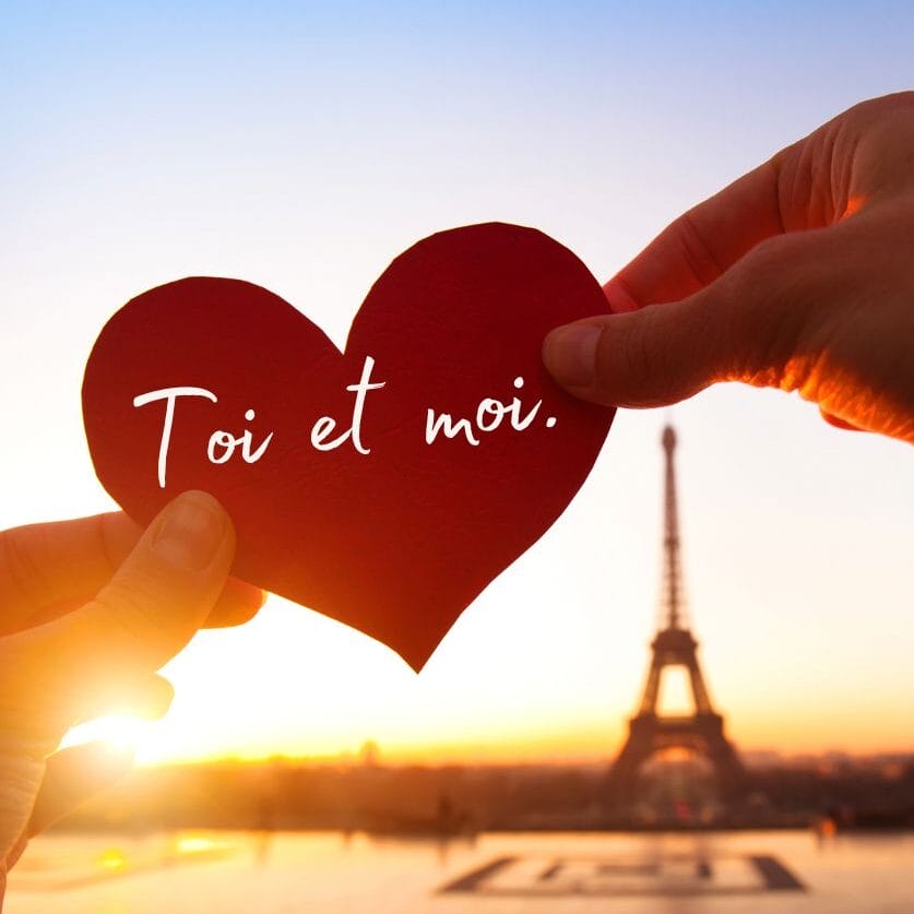 The image shows two hands holding a red heart-shaped cutout with the words "Toi et moi" (French for "You and me") written on it. In the background, the Eiffel Tower is visible at sunset, creating a romantic atmosphere with the warm light of the sky.