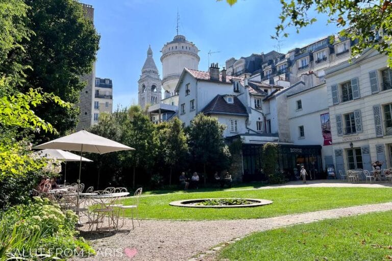 This image shows a peaceful outdoor setting at Cafรฉ Renoir in Montmartre, Paris. The cafรฉโs green glass exterior is covered with vines, and a chalkboard sign near the entrance welcomes guests. People are seated at tables in the garden, enjoying the sunny day, while others walk along the gravel paths. The surrounding area is lush with trees and greenery, creating a tranquil ambiance.