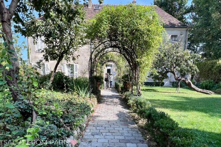 A picturesque garden pathway leading to the entrance of the Musรฉe de Montmartre in Paris, framed by lush green archways covered in climbing plants. The cobblestone path is surrounded by neatly trimmed hedges, flowering bushes, and a gracefully curved tree on the right. The historic building in the background, with its pale faรงade and shuttered windows, reflects the artistic and bohemian charm of Montmartre.