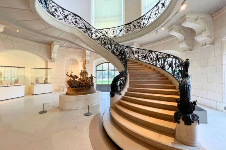 This image captures a grand, curved staircase at the Petit Palais, with intricate wrought iron railings and soft natural light streaming in through large windows. At the base of the staircase is a detailed sculpture, adding to the elegant ambiance of the space. The architecture features classical elements, including arches and ornate details, while a large mural can be seen overhead, just above the staircase.
