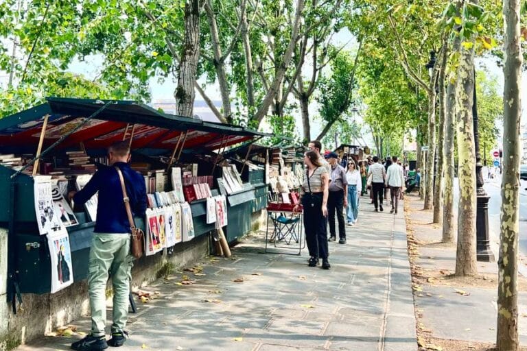 Bouquinistes along the seine with their green wooden boxes. Pedestrians are walking buy taking a look at the books. The trees are green and lined up the street