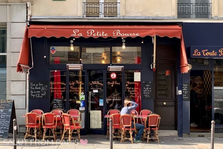 A charming Parisian cafรฉ with a red awning and matching red chairs arranged outside, inviting diners to enjoy the ambiance of the city street.