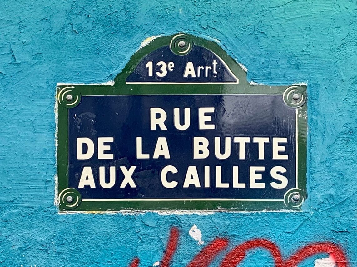 Street sign reading "Rue de la Butte aux Cailles" in the 13th arrondissement of Paris, mounted on a vibrant turquoise wall with graffiti accents.