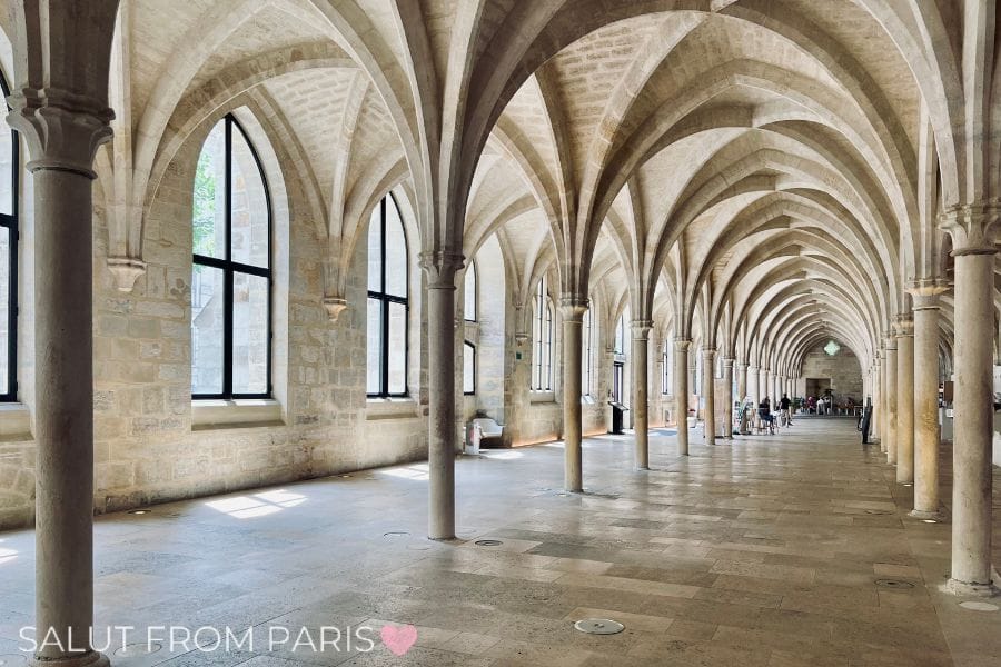 A long, vaulted corridor with Gothic arches inside the Collège des Bernardins in Paris. The sunlit stone interior features tall windows along one side, creating a serene and historic atmosphere.