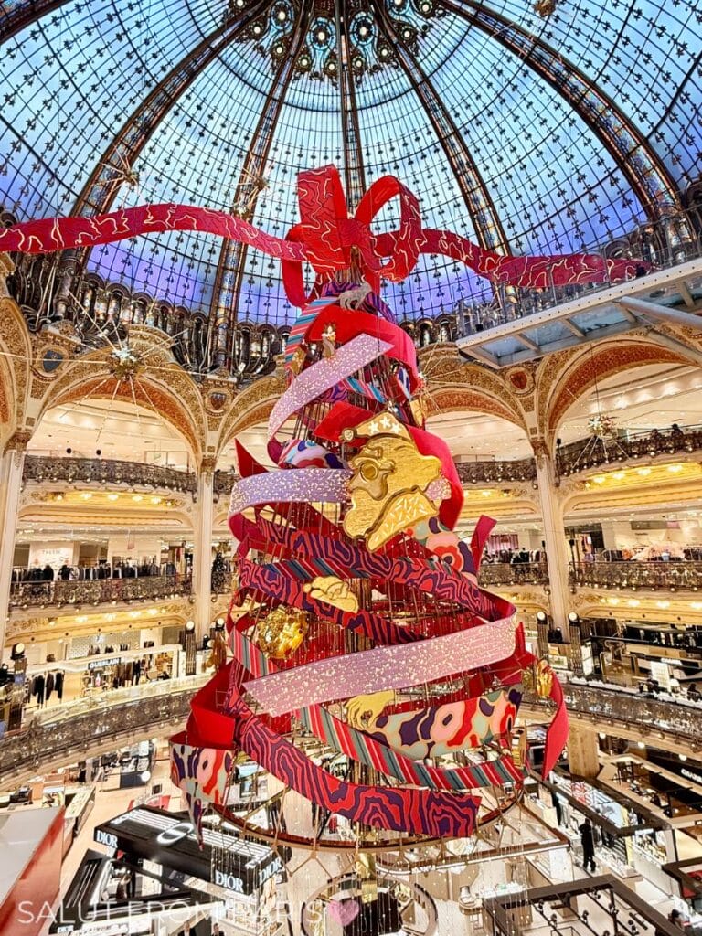 Christmas tree at Galeries Lafayette Haussmann 2025, designed by Jeanne Detallante, featuring swirling red and patterned ribbons, golden ornaments, and a giant bow, set under the department store’s iconic glass dome.