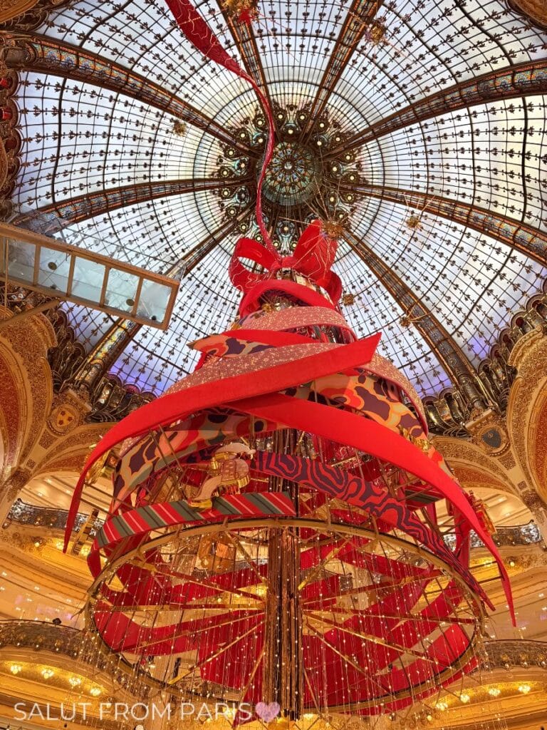 Christmas tree at Galeries Lafayette Haussmann 2025, designed by Jeanne Detallante, featuring swirling red and patterned ribbons, golden ornaments, and a giant bow, set under the department store’s iconic glass dome.