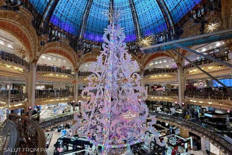 This image showcases the magnificent Christmas tree inside the Galeries Lafayette in Paris. The tree is adorned with intricate decorations and is set beneath the iconic glass dome, illuminated by blue and purple lights that create a magical holiday ambiance.