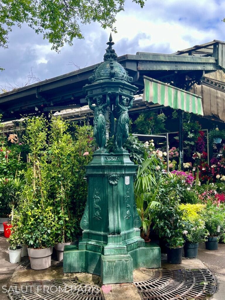 A classic green Wallace fountain in Paris, surrounded by lush potted plants outside a flower market. The fountain's ornate design adds charm to the vibrant setting of greenery and floral displays at the Marche aux Fleurs in Paris