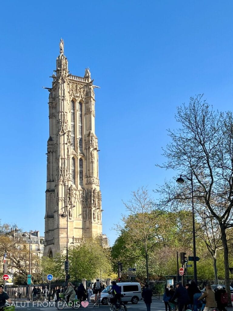 The Saint-Jacques Tower in Paris stands tall against a clear blue sky, showcasing its intricate Gothic architecture. Trees and pedestrians fill the area below, adding to the vibrant city scene.