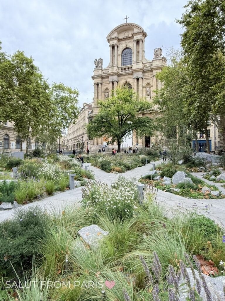 Landscaped garden with winding paths, ornamental grasses, and white flowering plants in front of the Church of Saint Paul Saint Louis in the Marais district of Paris. The historic stone church rises above the greenery beneath a cloudy sky while visitors walk through the square.