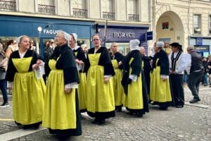 This image features a group of Brittany dancers dressed in traditional costumes, including black and yellow outfits with white lace accents. They are gathered in a cobblestone street, likely performing or participating in a cultural event, with buildings and shop signs visible in the background.
