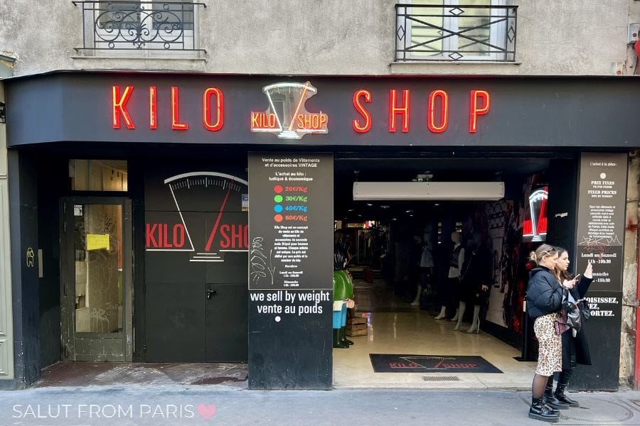 Front entrance of Kilo Shop, a vintage clothing store in Paris with bright red neon signage above the storefront. A sign at the entrance explains that clothing is sold by weight, and shoppers stand outside the open store.Image: uploaded image
Alt text: Cover image for a travel guide titled “Paris Without The Overwhelm” with the subtitle “The Calm Travel Guide.” The Eiffel Tower rises above the Paris skyline beneath dramatic gray clouds, with rooftops and apartment buildings filling the foreground.