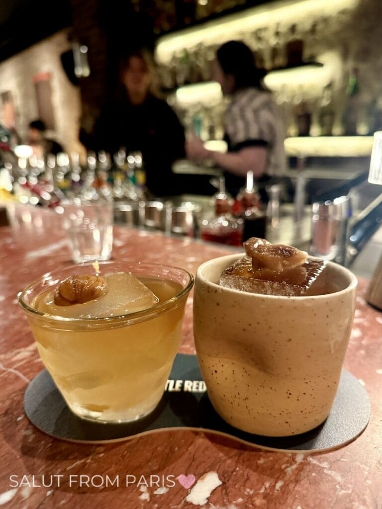 Two cocktails served on a marble bar in a dimly lit Paris cocktail lounge. One drink is presented in a glass and the other in a ceramic cup, both topped with large ice cubes and garnishes while bartenders work in the softly blurred background.