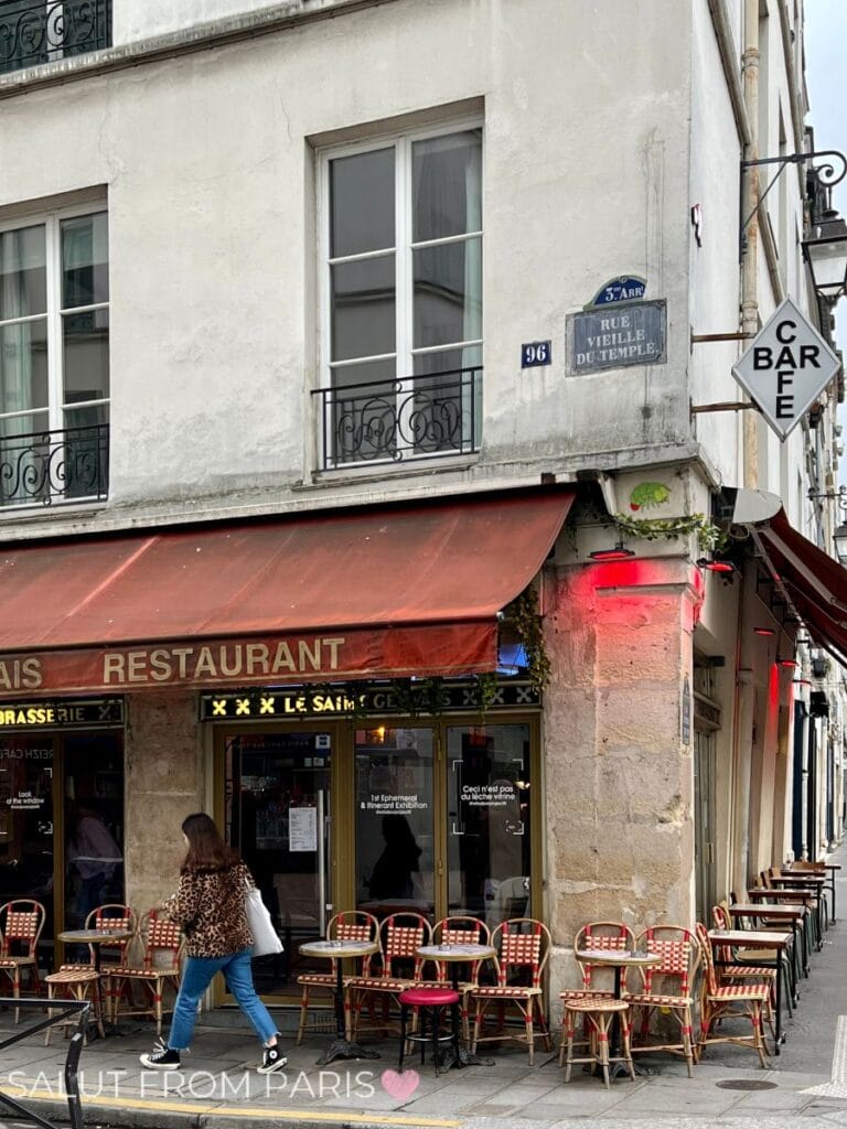 A cozy corner of Rue Vieille du Temple featuring a quaint Parisian restaurant. Red café chairs and tables spill onto the sidewalk, creating a classic French dining ambiance. The vintage building facades add timeless charm.