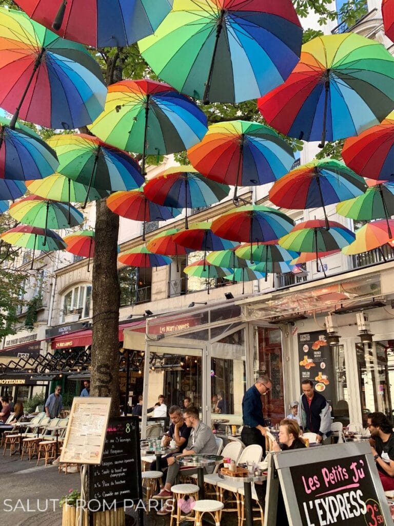 A lively café scene under colorful umbrellas in the Marais. Diners enjoy meals on a sunny terrace with vibrant rainbow decor. The playful umbrellas create a cheerful, inviting atmosphere.