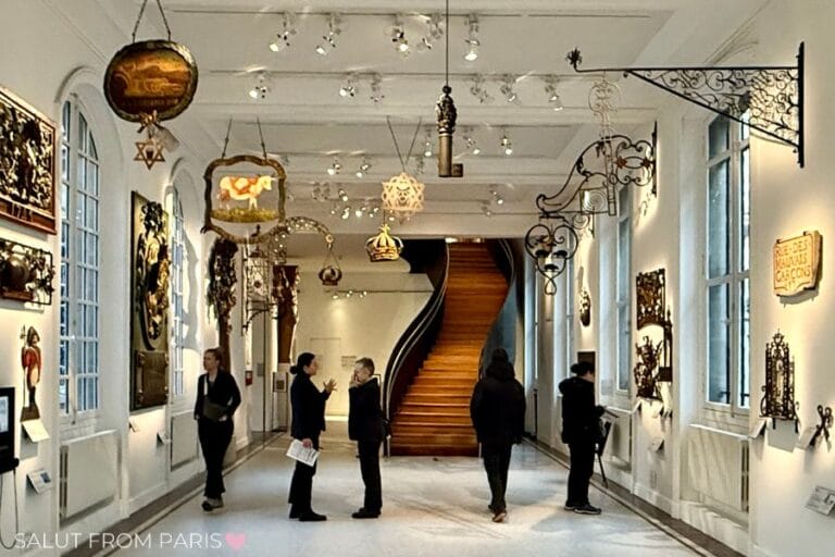 This image captures a hallway in the Musรฉe Carnavalet, Paris, featuring a collection of historical Parisian shop signs suspended from the ceiling and mounted on the walls. The space is well-lit with natural light streaming through large windows, and visitors are seen admiring the exhibits. A striking wooden spiral staircase in the background adds an elegant architectural touch.