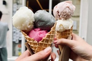Two hands hold waffle cones filled with colorful scoops of ice cream in front of an ice cream shop. The cones feature flavors like white coconut, deep pink raspberry, dark gray sesame, and soft pink rose. The blurred background shows a minimalist storefront with a cozy ambiance.