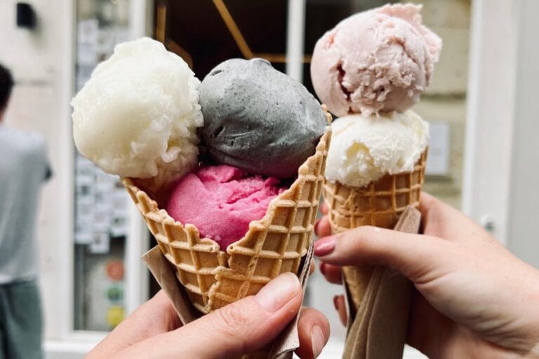 Two hands hold waffle cones filled with colorful scoops of ice cream in front of an ice cream shop. The cones feature flavors like white coconut, deep pink raspberry, dark gray sesame, and soft pink rose. The blurred background shows a minimalist storefront with a cozy ambiance.