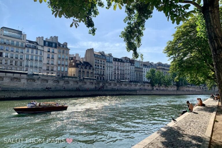 People sitting quietly along the shaded Seine riverbank in central Paris, with a wooden boat passing by.