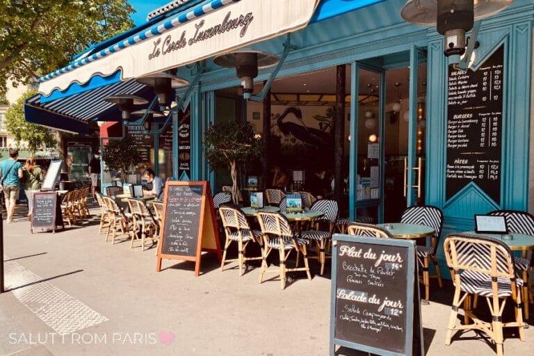 Street view of Cafรฉ Le Cercle Luxembourg in Paris with typical striped chairs, awnings, and chalkboard menus in the sun.