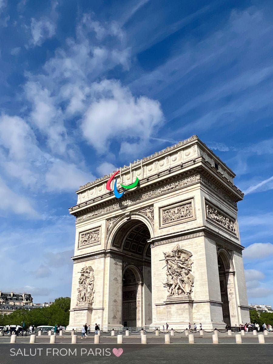The Arc de Triomphe in Paris stands tall under a vivid blue sky with scattered clouds, adorned with the colorful 2024 Paralympic Games symbol in red, blue, and green. The grand monument is detailed with sculptural reliefs and surrounded by tourists at its base. “SALUT FROM PARIS” with a pink heart is watermarked at the bottom.