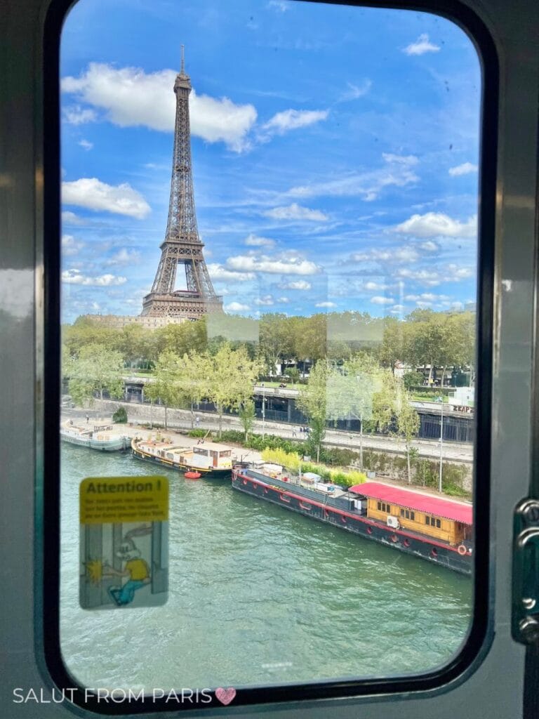 The Eiffel Tower rises against a bright blue sky filled with scattered clouds, seen through a Metro train window crossing the Seine River. Houseboats are moored along the riverside below, and a cartoon warning sticker with "Attention!" is posted on the window. 