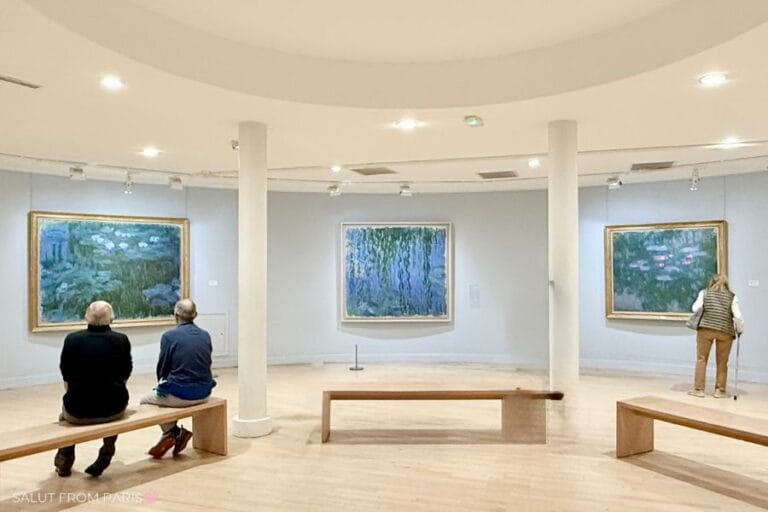 Monet Water Lilies Gallery Room Wide-angle view of a softly lit circular gallery with large Monet water lily paintings displayed on curved walls; visitors sit on minimalist benches and admire the art.