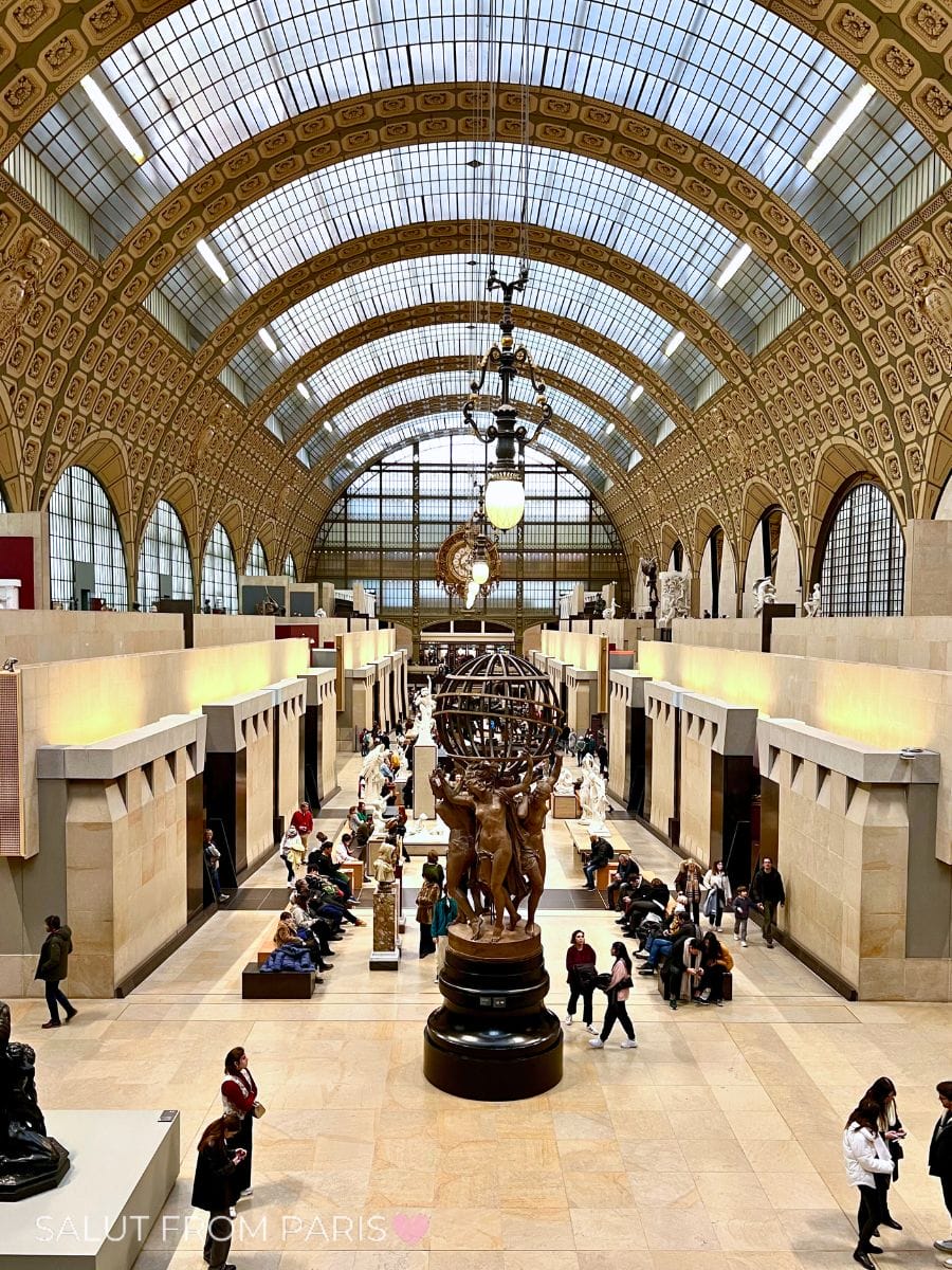 The grand central hall of the Musée d'Orsay in Paris, with its soaring arched glass ceiling and ornate gold-trimmed architecture, filled with sculptures and museum visitors. A bronze statue of nude figures holding up an armillary sphere stands prominently in the center, surrounded by seated and strolling guests