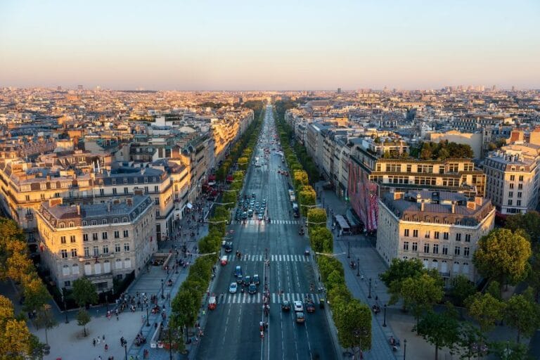 A golden-hour aerial view of the Champs-รlysรฉes in Paris, with rows of elegant Haussmann-style buildings and tree-lined sidewalks stretching into the distance. The broad avenue is busy with cars, buses, and pedestrians as the soft evening sunlight casts a warm glow across the city.
