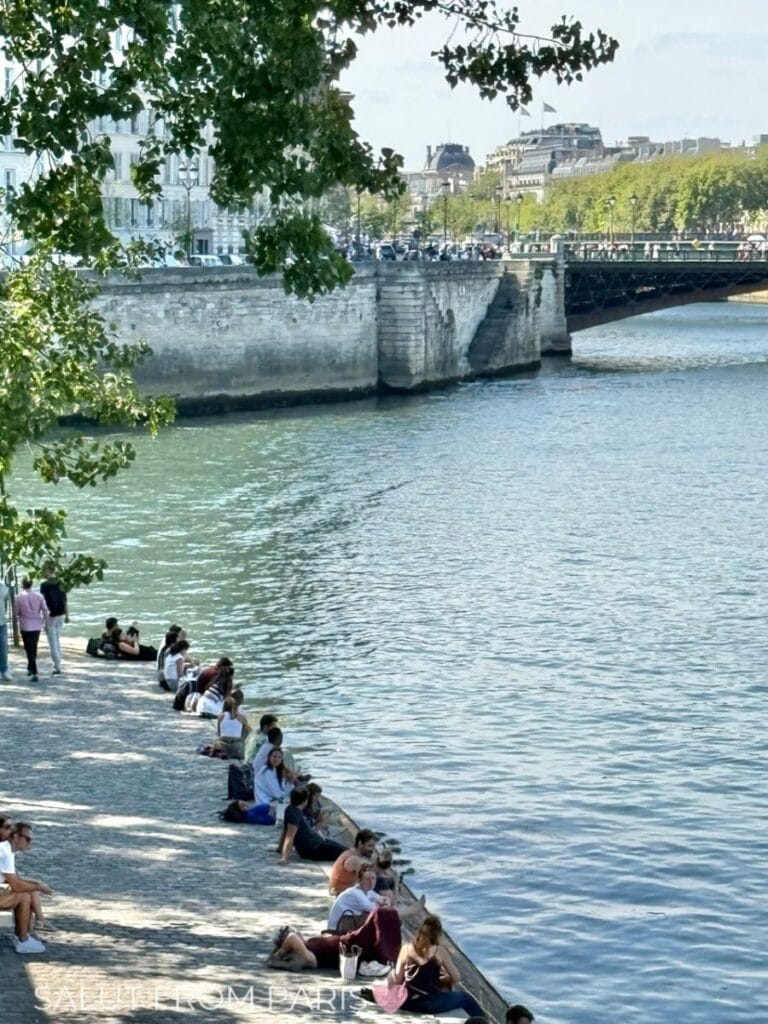 People relaxing along the Seine River on a sunny day in Paris in May, with leafy trees framing the scene and a stone bridge and historic buildings in the background.