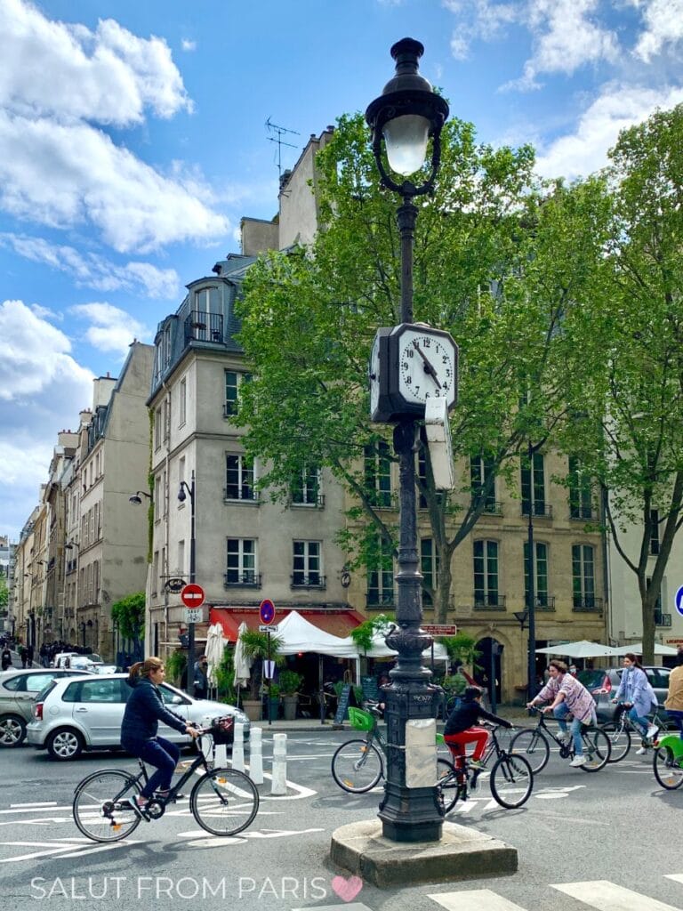 Cyclists ride through a Paris street in May past a vintage lamppost with a clock, surrounded by leafy trees, classic buildings, and a small cafรฉ on a bright day.