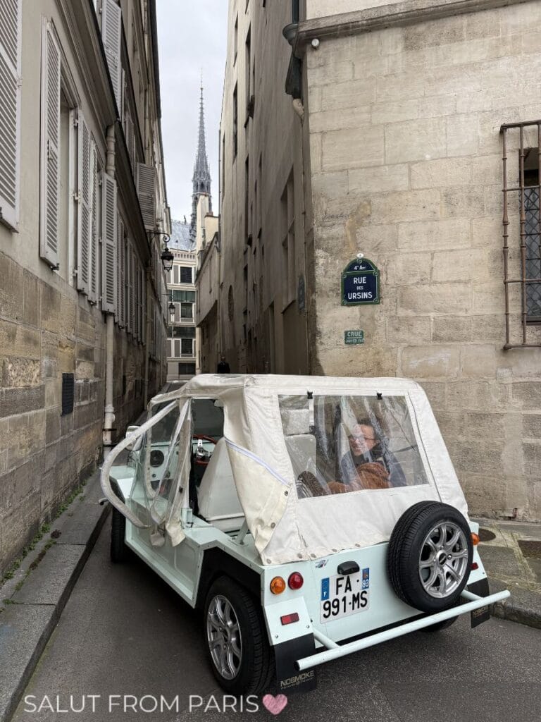 A small open car parked in a narrow Paris street in May near a sign reading "Rue des Ursins," with stone buildings and a church spire rising in the background.